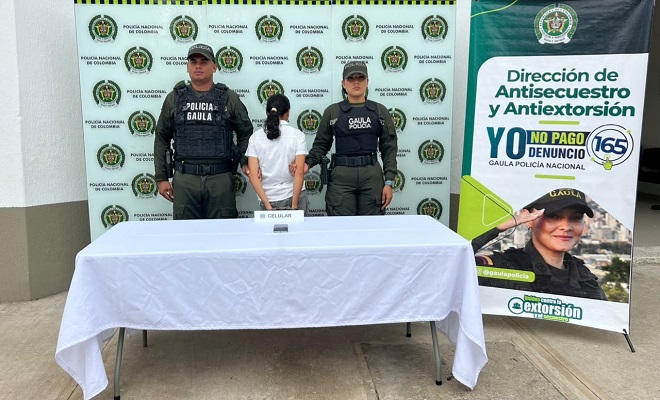 Un celular incautado sobre una mesa con mantel blanco, una persona de sexo femenino capturada por el delito de extorsi&oacute;n, acompa&ntilde;ada de dos uniformados del GAULA de la Polic&iacute;a Nacional brindando seguridad.