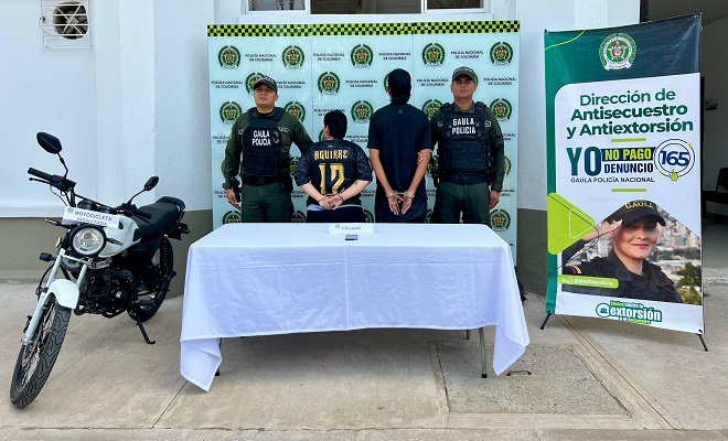 Una motocicleta incautada, un celular incautado sobre una mesa con mantel blanco, dos personas de espaldas capturadas por el delito de extorsi&oacute;n en compa&ntilde;&iacute;a de dos funcionarios del GAULA de la Polic&iacute;a Nacional brindando seguridad. 
