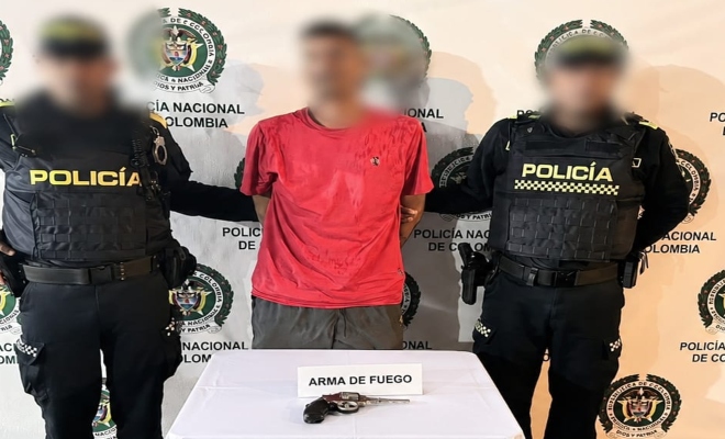 Dos funcionarios policiales con un ciudadano capturado frente a pendón institucional frente a mesa con arma de fuego incautada