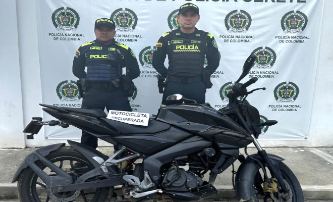 Dos funcionarios policiales con una  motocicleta frente a pendón institucional 