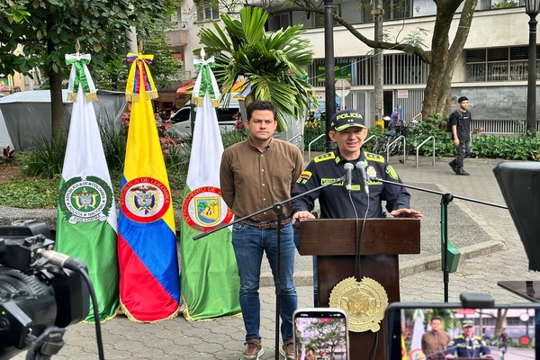 Via p&uacute;blica, tres banderas en hasta, Polic&iacute;a Nacional, Colombia, Medell&iacute;n frente a ellas, dos personas, un polic&iacute;a y un ciudadano, las c&aacute;maras de video en rueda de prensa.   