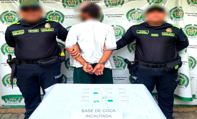 Dos funcionarios policiales con un ciudadano capturado frente a pendón institucional frente a ellos material incautado sobre una mesa
