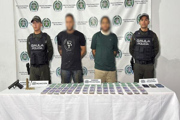 Un pendón con escudo de la Policía Nacional, en pie cuatro personas dos uniformadas, una mesa con elementos, dinero, un arma y munición