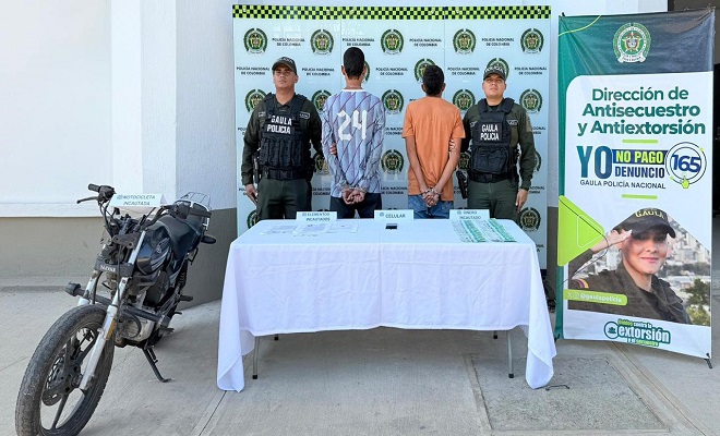 Dos personas de espalda capturadas por el delito de extorsi&oacute;n, elementos incautados sobre una mesa con mantel blanco, dos funcionarios de la Polic&iacute;a brindando seguridad y una motocicleta incautada.
