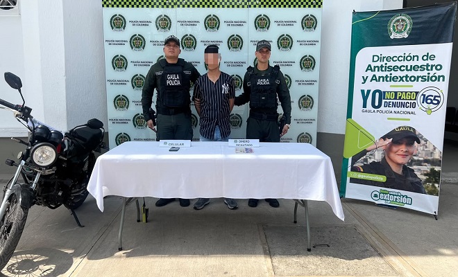 Una persona capturada por el delito de extorsi&oacute;n junto a una mesa con mantel blanco donde est&aacute;n ubicados los elementos incautados, al lado una motocicleta incautada y dos funcionarios del grupo GAULA de la Polic&iacute;a Nacional brindando seguridad