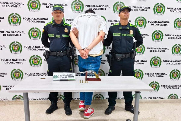 Hombre capturado de camiseta blanca en medio de dos policías, en frente una mesa y sobre ella el arma incautada