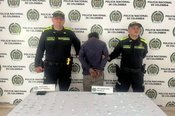 Hombre capturado de espalda, vistede buo azul y pantalon gris en medio de dos policías, en frente se encuentra una mesa con material incautado