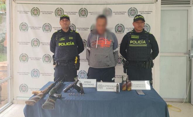 Tres personas de pie frente a un fondo institucional de la Policía Nacional; en primer plano hay una mesa con armas largas, munición y otros objetos exhibidos de forma organizada.