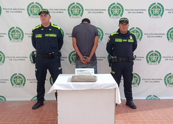 Hombre capturado en medio de dos policias, en frente una mesa y sobre ella marihuana incautada