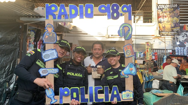 Agentes de la Polic&iacute;a comparten con un ciudadano en un mercado, utilizando un marco decorado con el logo de la emisora, promoviendo el acercamiento y la comunicaci&oacute;n entre la instituci&oacute;n y la comunidad