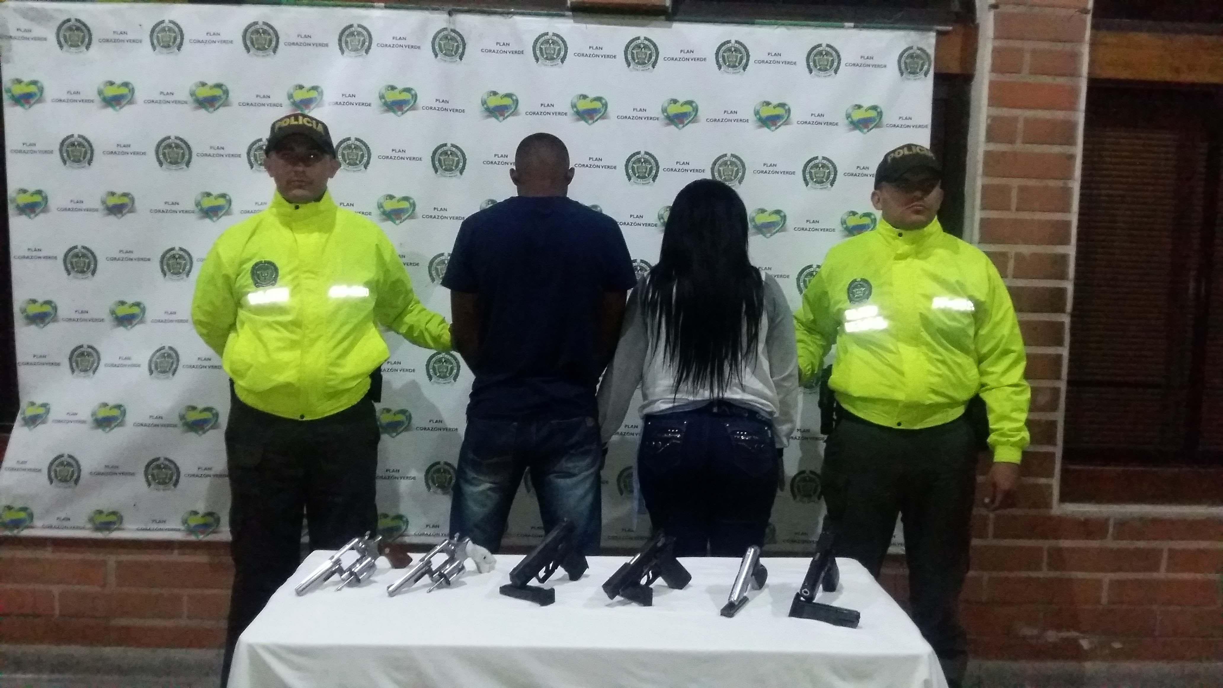 Dos personas de espaldas detrás de seis armas de fuego incautadas son custodiadas por dos policías 