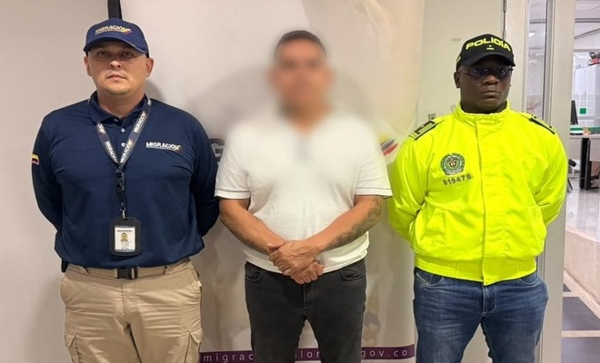 Tres personas de pie en un espacio interior frente a un panel informativo; a los lados se observan una persona con uniforme de Migraci&oacute;n Colombia y otra con uniforme de la Polic&iacute;a Nacional, mientras la persona ubicada en el centro viste camiseta clara y pantal&oacute;n oscuro.