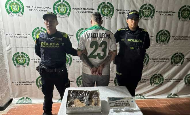 Hombre capturado en medio de dos Polic&iacute;as, en frente armas de fuego incautadas
