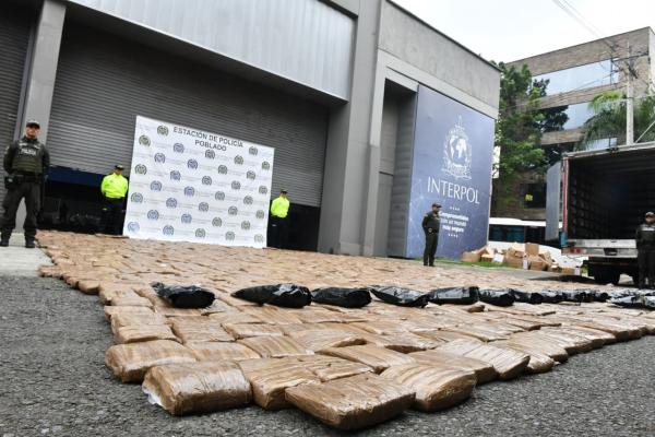 Se observa una gran cantidad de paquetes rectangulares envueltos en plástico, en el suelo como resultado de una incautación de marihuana. Al fondo se ven uniformados de la Policía Nacional, un camión y un banner institucional.   