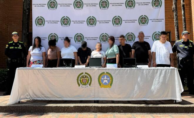 Grupo de personas de pie detrás de una mesa con varios computadores portátiles y avisos informativos, frente a un fondo institucional de la Policía Nacional de Colombia; dos personas uniformadas se encuentran en los extremos de la fila.