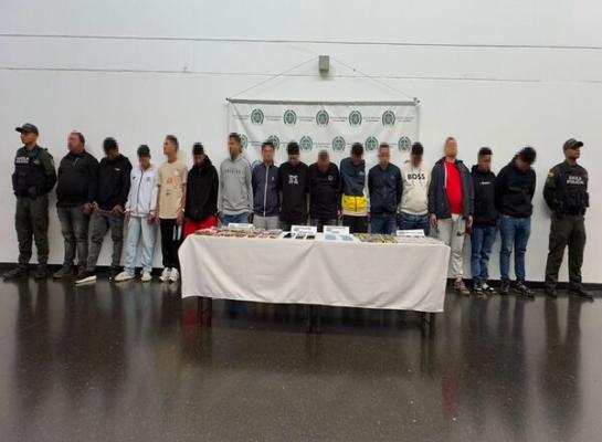 Espacio interior con fondo institucional de la Polic&iacute;a Nacional de Colombia donde una fila de varias personas est&aacute; de pie frente a una mesa cubierta con un mantel blanco; sobre la mesa hay objetos organizados en filas y dos personas uniformadas se ubican en los extremos.