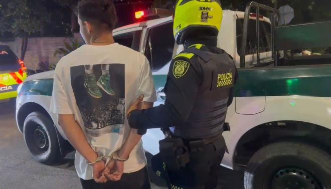 hombre capturado de camiseta blanca camina junto a un polic&iacute;a hacia una patrulla