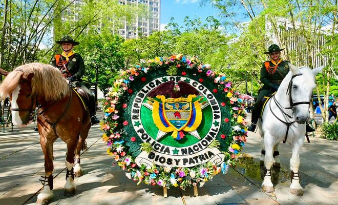 Estructura floral circular con el escudo Polic&iacute;a Nacional ubicada entre dos caballos montados por personal uniformado en un espacio p&uacute;blico arborizado.